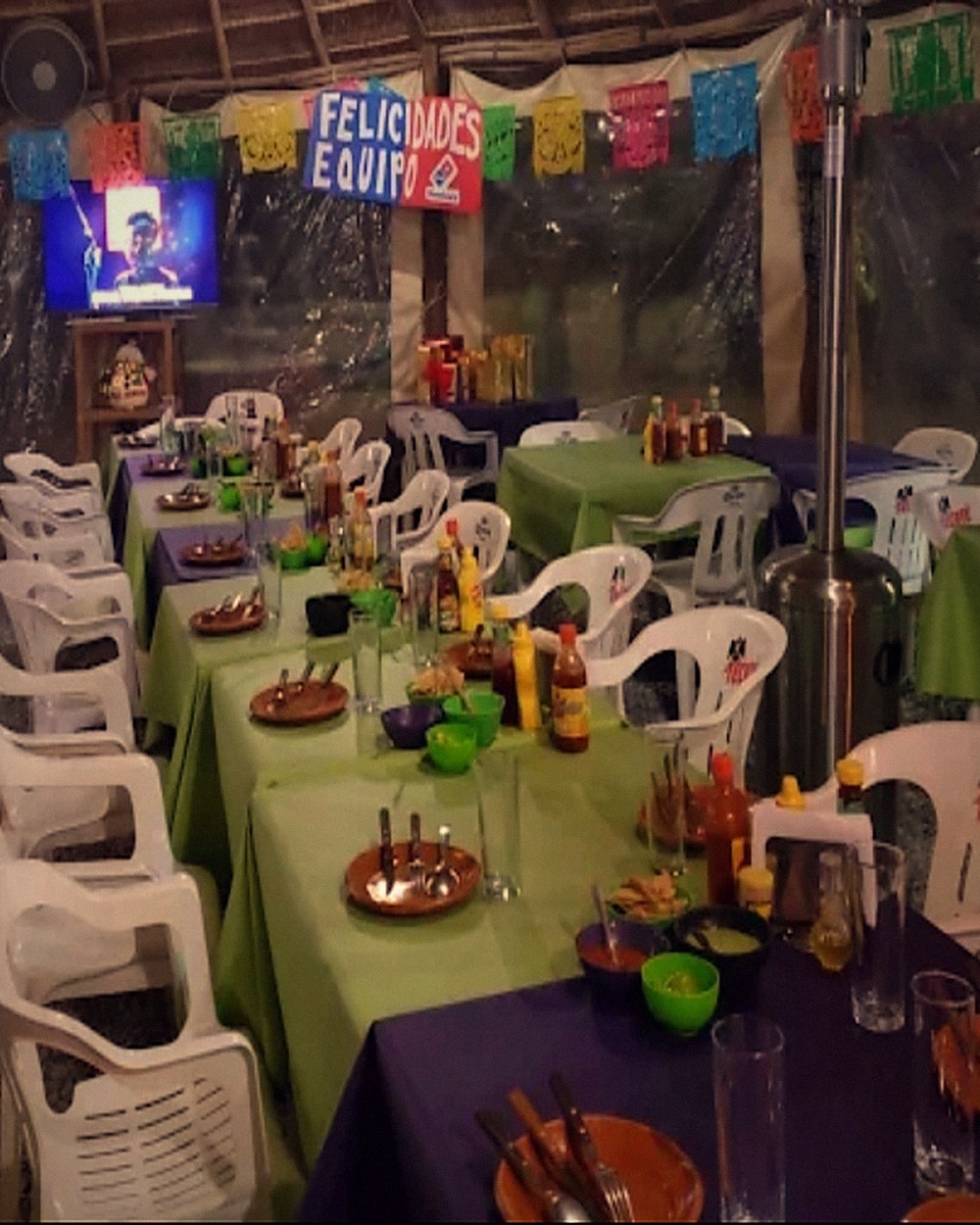 Mesas largas servidas de noche bajo la palapa, con papel picado y calentadores